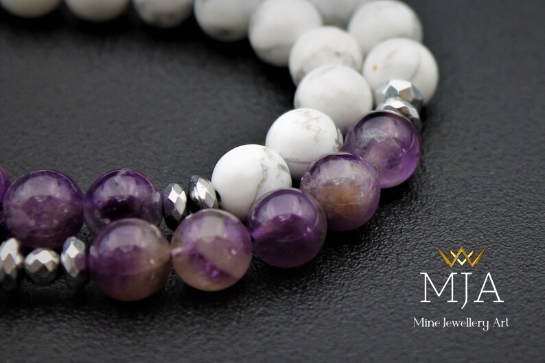 K&ouml;nnte beinhalten: Nahaufnahme eines Armbands aus wei&szlig;en und violetten Perlen. Das Armband besteht aus wei&szlig;en Howlith-Perlen und Amethyst-Perlen. Das Armband liegt auf einer schwarzen Oberfl&auml;che.