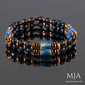May include: A double-strand bracelet with black beads, copper spacers, and a blue lapis lazuli focal bead. The bracelet is on a black reflective surface. "MJA Mine Jewellery Art" is printed in white on a black background.