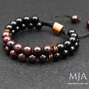 May include: A double-strand bracelet featuring alternating rows of black and burgundy-colored beads. The bracelet has a sliding knot closure with copper-colored accents and small, dark beads at the ends. The jewelry is displayed on a dark surface.
