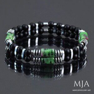 Op de afbeelding: Een dubbele armband met zwarte onyx kralen, zilveren tussenkralen en een groene en roze edelsteen. De armband ligt op een reflecterend oppervlak. "MJA Mine Jewellery Art" is afgedrukt in de rechterbenedenhoek van de afbeelding.