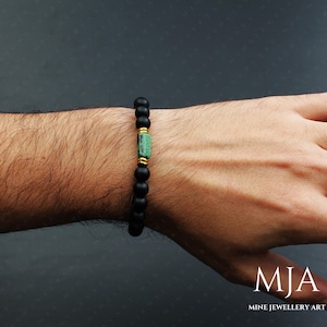 May include: Black beaded bracelet with a green and gold gemstone accent. The bracelet is on a person's wrist.