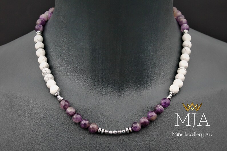 K&ouml;nnte beinhalten: Eine violette Amethyst- und wei&szlig;e Howlith-Perlenkette mit silbernen Akzenten. Die Kette befindet sich an einem Mannequin-Hals. Der Text "MJA Mine Jewellery Art" befindet sich in der unteren rechten Ecke des Bildes.