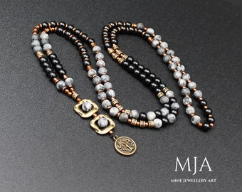 May include: A layered necklace with black and grey beads, copper accents, and a gold pendant with a tree design. The necklace has two square-shaped charms with grey and black stones.