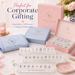 May include: A mahjong set featuring clear acrylic tiles with colorful symbols, three clear acrylic tile racks with the text YOUR BRAND, a light blue fabric mat, a pink storage box, and a pink tray, all branded with the YOUR BRAND logo.