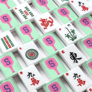 May include: Close-up of a set of white Mahjong tiles. The tiles feature various symbols, numbers, and characters in red, green, and black. Some tiles have a pink and blue design. The tiles are arranged in a grid.