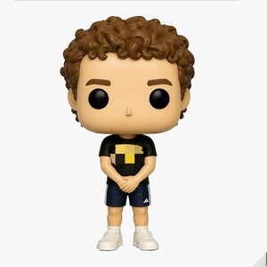 May include: A vinyl figure of a person with brown curly hair, black eyes, and a tan complexion. The figure wears a black t-shirt with a yellow and black design, navy blue shorts, white socks, and white sneakers. The figure is standing.
