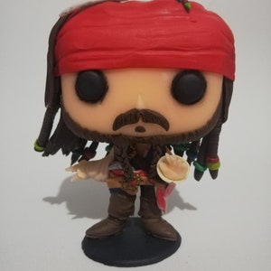 May include: A Funko Pop! figure of Captain Jack Sparrow from the Pirates of the Caribbean film series. The figure features a red bandana, braided hair with colorful beads, and a brown outfit. It stands on a black base.