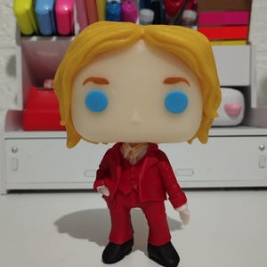 Pode incluir: Figura de vinil Funko Pop de um personagem com cabelo loiro, olhos azuis e um fato vermelho. A figura está em pé, com sapatos pretos e luvas brancas. A figura é um item colecionável.