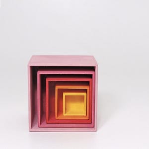 May include: A set of four nested square open boxes arranged in a gradient color scheme. The largest outer box is light pink, followed by a medium pink box, a red box, and the smallest innermost box which is a bright yellow.