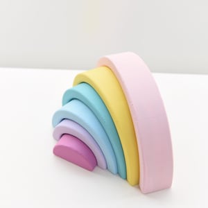 Puede incluir: Un conjunto de cinco arcos de madera encajables en degradado de arcoíris. Los colores incluyen un arco grande rosa pálido, seguido de amarillo, turquesa, azul claro, lavanda y un arco pequeño magenta. Cada pieza tiene un acabado mate suave.