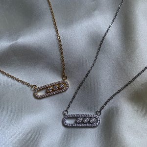 May include: Two necklaces with pendant designs. One necklace has a gold chain and pendant, the other has a silver chain and pendant. Each pendant is an oval shape with three round stones in the center and smaller stones around the oval.