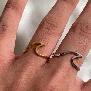 May include: Two metal rings worn on adjacent fingers, each featuring a stylised wave design. One ring has a polished gold-tone finish, while the other ring has a polished silver-tone finish. Both bands are thin and follow the shape of the wave.