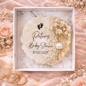 May include: A baby shower keepsake in a white box. The centerpiece is a cream-coloured, resin-like coaster with gold accents, baby footprints, and the text "Peters Baby Shower 10/02/2024." It is decorated with dried flowers and a pearl.