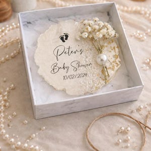 May include: A baby shower gift box containing a decorative paper insert. The insert is inscribed with "Peter's Baby Shower" and the date 10/02/2024, plus a footprint graphic. A small bouquet of white flowers and a pearl embellishment are also present.