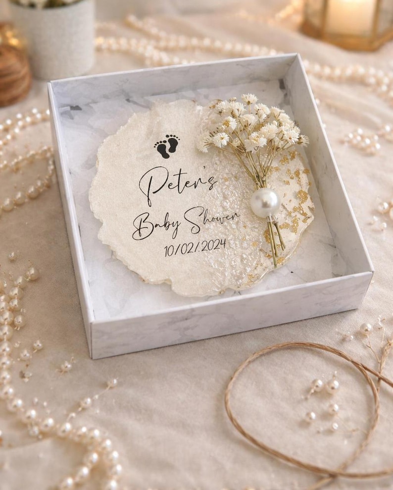 May include: A baby shower gift box containing a decorative paper insert. The insert is inscribed with "Peter's Baby Shower" and the date 10/02/2024, plus a footprint graphic. A small bouquet of white flowers and a pearl embellishment are also present.