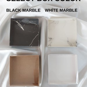 May include: Four square boxes in different colours: black marble, white marble, brown, and white. The text "SELECT BOX COLOUR" is at the top, with the colour options below. The boxes are on a white, silky surface.