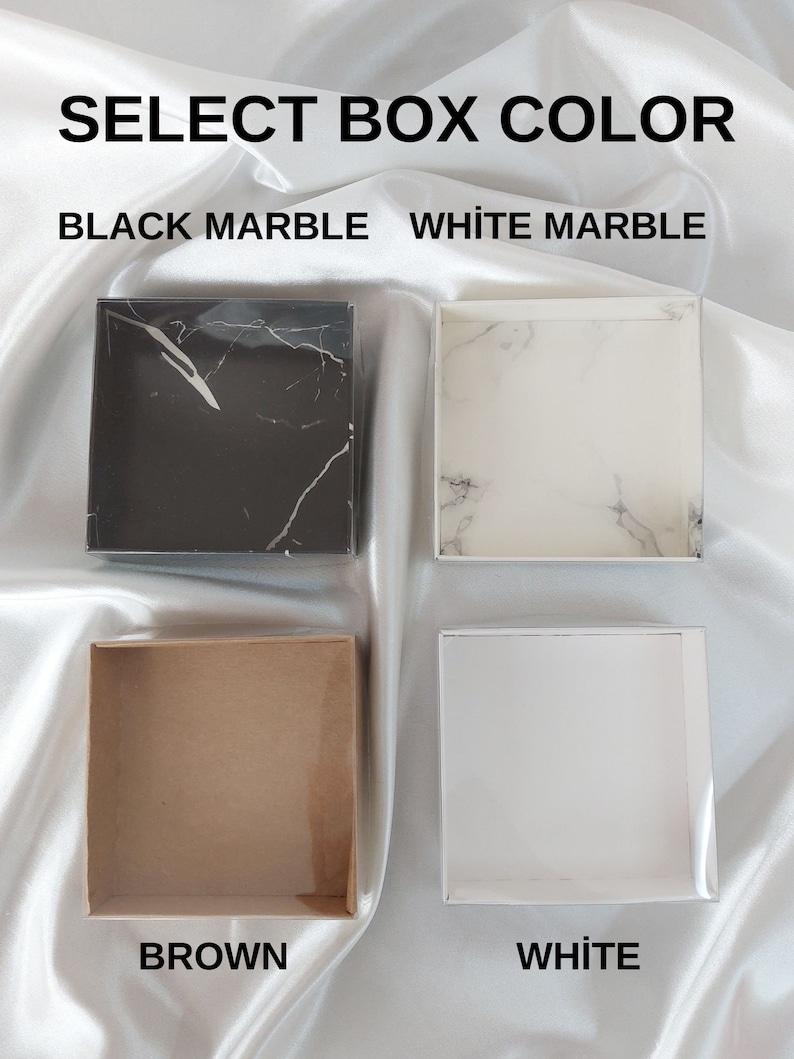 May include: Four square boxes in different colours: black marble, white marble, brown, and white. The text "SELECT BOX COLOUR" is at the top, with the colour options below. The boxes are on a white, silky surface.