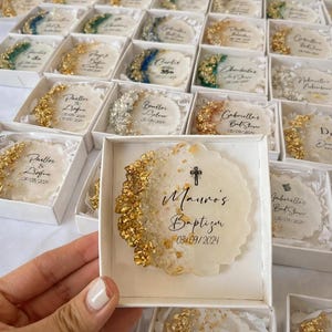 May include: A selection of personalised favours in white boxes. Each favour is a decorative, irregularly shaped coaster with gold leaf accents and custom text. The text includes names, dates, and event details, suitable for a baptism or similar celebration.