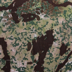 May include: A close-up of a camouflage fabric with a brown, green, and beige pattern. The fabric has a textured surface and a slightly faded appearance.