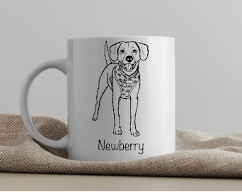 May include: White ceramic mug with a black line drawing of a dog wearing a patterned harness. The dog is looking to the right and has its tongue sticking out. The text "Newberry" is printed below the dog.