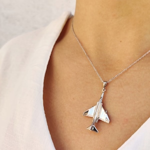 Pode incluir: Um colar de prata com um pingente de avião detalhado. O pingente é uma réplica de um caça.