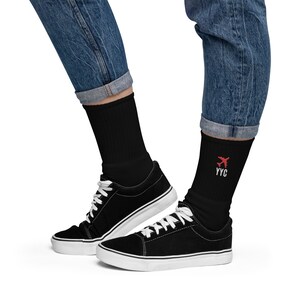 May include: A pair of black socks with a red airplane and the text "YYC" embroidered on one sock. The socks are being worn with black trainers and blue jeans.