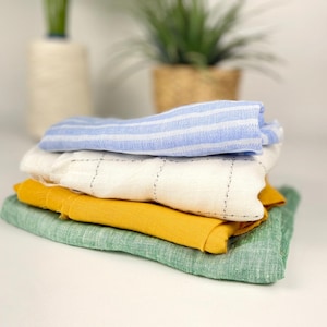Può includere: Una pila di panni di lino piegati in vari colori: azzurro a righe, bianco con motivo a griglia, giallo senape e verde. I panni sono ben disposti, mostrando le loro trame e tonalità.