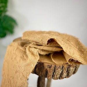 May include: A tan textured fabric draped over a wooden log. The fabric has a frayed edge and a soft, wrinkled texture.