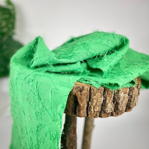 Könnte beinhalten: Ein leuchtend grüner Stoff mit einer strukturierten, ausgefransten Oberfläche. Der Stoff ist über einen Holzstamm gelegt.