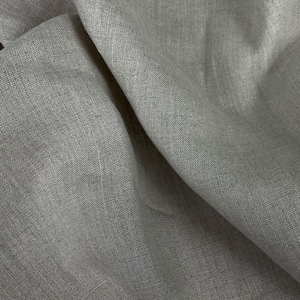 Può includere: Primo piano di un tessuto grigio chiaro con una texture sottile. Il tessuto è piegato e drappeggiato, mostrando la sua morbidezza e la sua caduta.