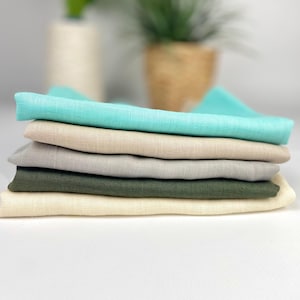 Può includere: Una pila di sei tovaglioli di lino piegati in vari colori. Il tovagliolo superiore è turchese brillante, seguito da beige, grigio, verde oliva e crema. I tovaglioli sono ordinatamente impilati, mostrando il loro tessuto strutturato.