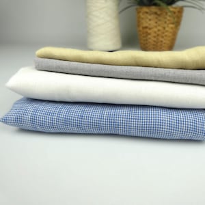 Può includere: Una pila di tessuti piegati in vari colori. Lo strato superiore è giallo chiaro, seguito da grigio, bianco e un motivo a scacchi blu e bianco. I tessuti sono ben disposti, suggerendo un tema tessile o artigianale.