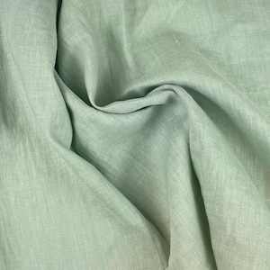 Può includere: Primo piano di un tessuto di lino verde chiaro con trama e pieghe visibili. Il tessuto ha un colore morbido e attenuato e sembra leggero. L'immagine evidenzia la trama naturale e il drappeggio del materiale, adatto per abbigliamento o arredamento.