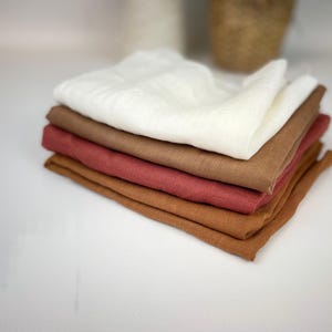 Può includere: Una pila di panni di lino piegati in tonalità di bianco, marrone e rosso-arancio. I panni sono ordinatamente disposti, mostrando i loro diversi colori e trame. L'immagine è ben illuminata, evidenziando i toni naturali del tessuto.