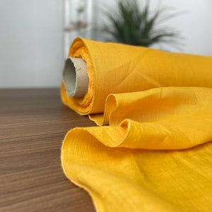 Può includere: Un rotolo di tessuto giallo brillante, srotolato su una superficie di legno. Il tessuto è morbido e ha un aspetto leggermente strutturato.