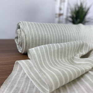 Può includere: Un rotolo di tessuto di lino naturale a righe bianche. Il tessuto è piegato e drappeggiato su una superficie di legno.