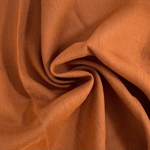 Può includere: Primo piano di un tessuto di lino color ruggine con una trama testurizzata. Il tessuto è piegato e drappeggiato, mostrando la sua texture morbida e naturale.