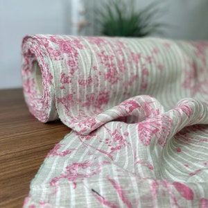 Può includere: Un rotolo di tessuto di lino bianco con strisce floreali rosa. Il tessuto è morbido e ha una lucentezza delicata.