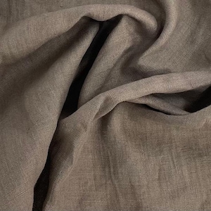 Può includere: Primo piano di un tessuto di lino marrone con una trama testurizzata. Il tessuto è piegato e drappeggiato, mostrando la sua texture morbida e naturale.