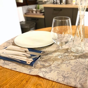 Op de afbeelding: Een wit dinerbord, een blauw-wit servet, een zilveren vork en mes, een helder wijnglas en een heldere glazen vaas met gedroogde witte bloemen op een beige placemat met een bloemenpatroon.