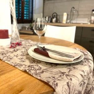 Op de afbeelding: Een close-up van een tafeldekking met een wit bord, een bordeauxrode servet en zilveren bestek. De tafel is bedekt met een beige linnen tafelkleed met een bloemenpatroon.