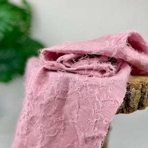Könnte beinhalten: Rosafarbener Stoff mit einer strukturierten, ausgefransten Oberfläche. Der Stoff ist gefaltet und über eine Holzoberfläche gelegt.