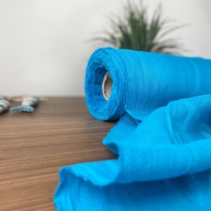 Può includere: Un rotolo di tessuto blu turchese brillante su una superficie di legno. Il tessuto è morbido e fluido, e ha un aspetto leggermente strutturato.