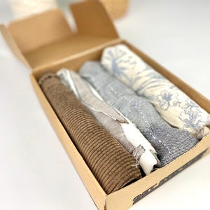 Peut inclure: Une boîte en carton marron avec un couvercle ouvert, révélant quatre carrés de tissu. Les carrés sont dans différentes nuances de bleu et de blanc, avec un carré présentant un motif floral. Les carrés de tissu sont pliés soigneusement à l'intérieur de la boîte.