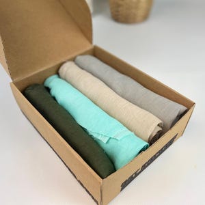 Könnte beinhalten: Eine offene Pappschachtel mit vier gerollten Leinen-Schals in Grün-, Türkis-, Beige- und Grautönen. Die Schals sind in der Schachtel angeordnet und zeigen ihre weiche Textur und lebendigen Farben.