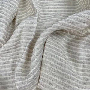 Puede incluir: Primer plano de una tela de rayas blancas y grises con un tejido texturizado. La tela está doblada y drapeada, mostrando la textura suave y natural.