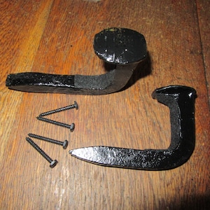 May include: Two black metal hooks with a nail head design. The hooks are attached to a wooden surface with screws.