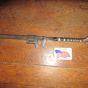 May include: A handcrafted metal adjustable wrench with a twisted handle and a looped end. The wrench is made of dark metal and rests on a wooden surface. A sticker with the American flag and the words "Proudly Made in USA" is also present.