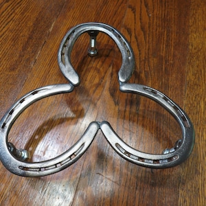 Puede incluir: Un salvamanteles decorativo de metal con forma de herradura. El salvamanteles está hecho de tres herraduras unidas con pernos. El metal tiene un acabado plateado. El salvamanteles está diseñado para sostener ollas y sartenes calientes.