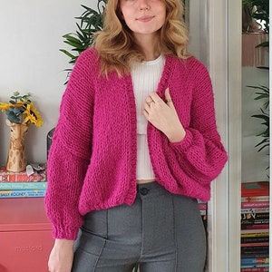 Op de afbeelding: Een felroze gebreide cardigan met lange mouwen en een ontspannen pasvorm. De cardigan is open aan de voorkant en heeft een licht getailleerde lengte.